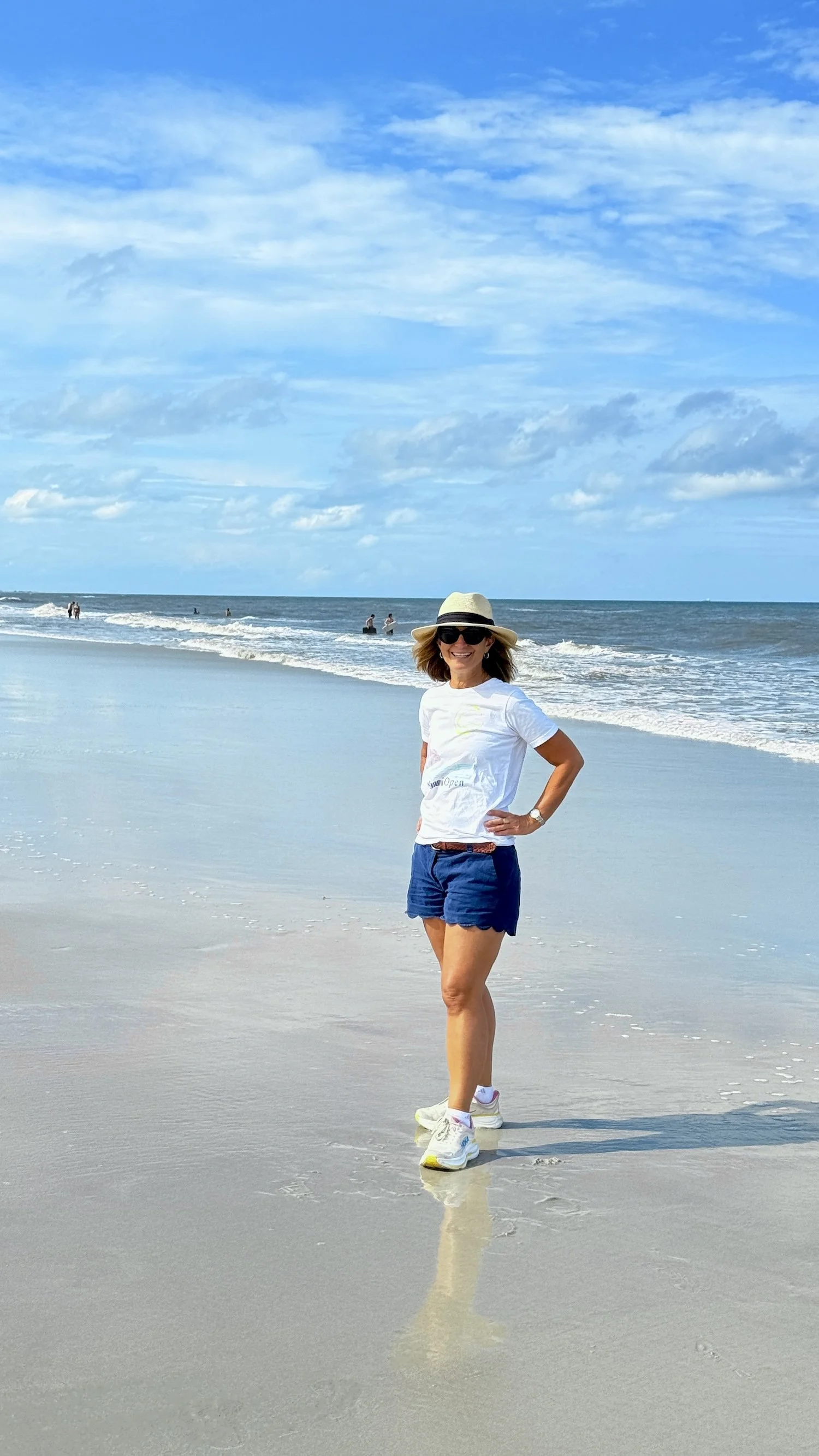A woman standing on a beach with her hands on her hips, wearing a hat, sunglasses, a white T-shirt, blue shorts, and sneakers, with the ocean and a partly cloudy sky in the background.