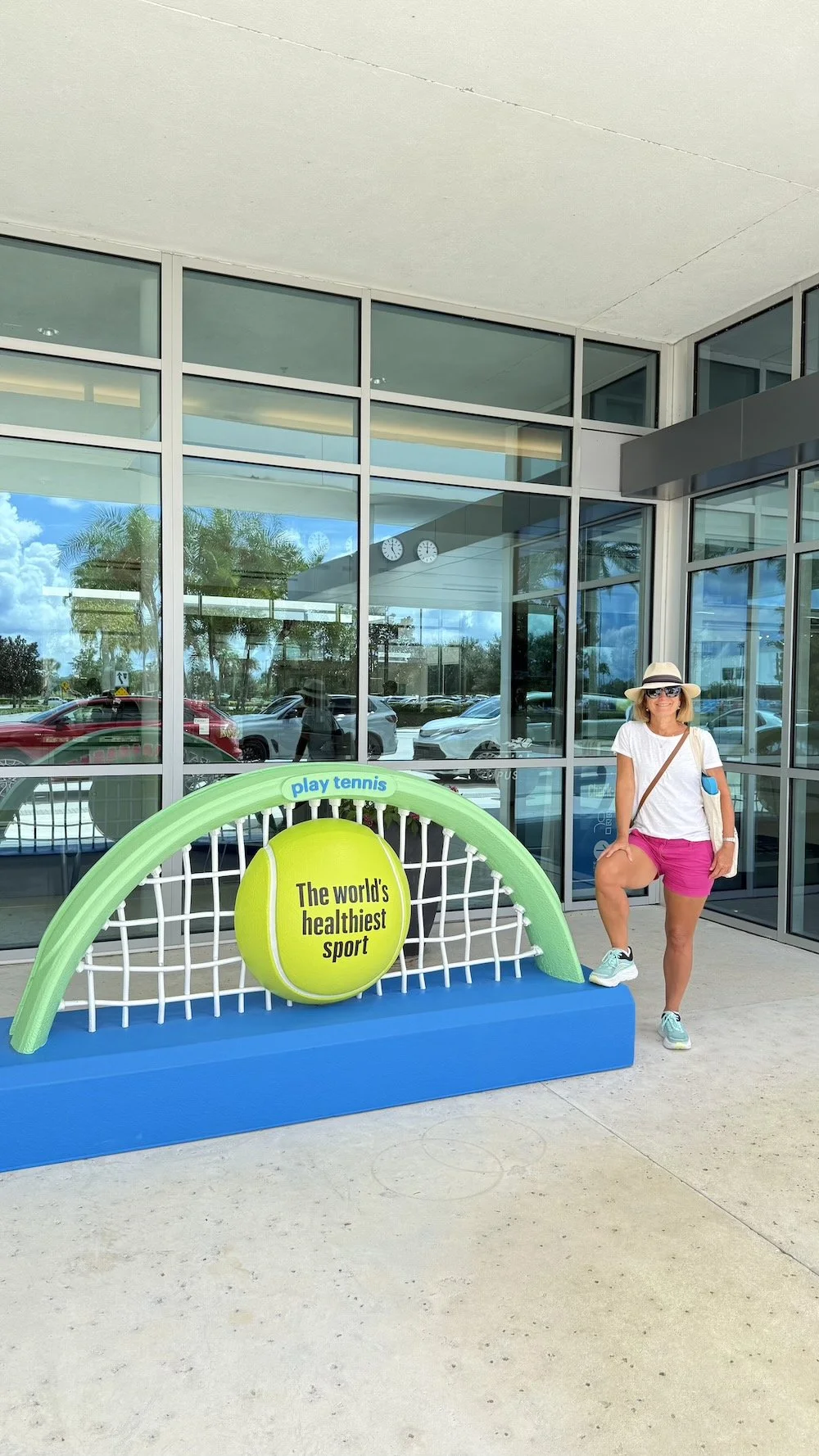 Woman in sunglasses, white t-shirt, pink shorts, and hat standing next to a tennis-themed display outside a modern glass building, with cars in the background and a large yellow tennis ball that reads "The world's healthiest sport."