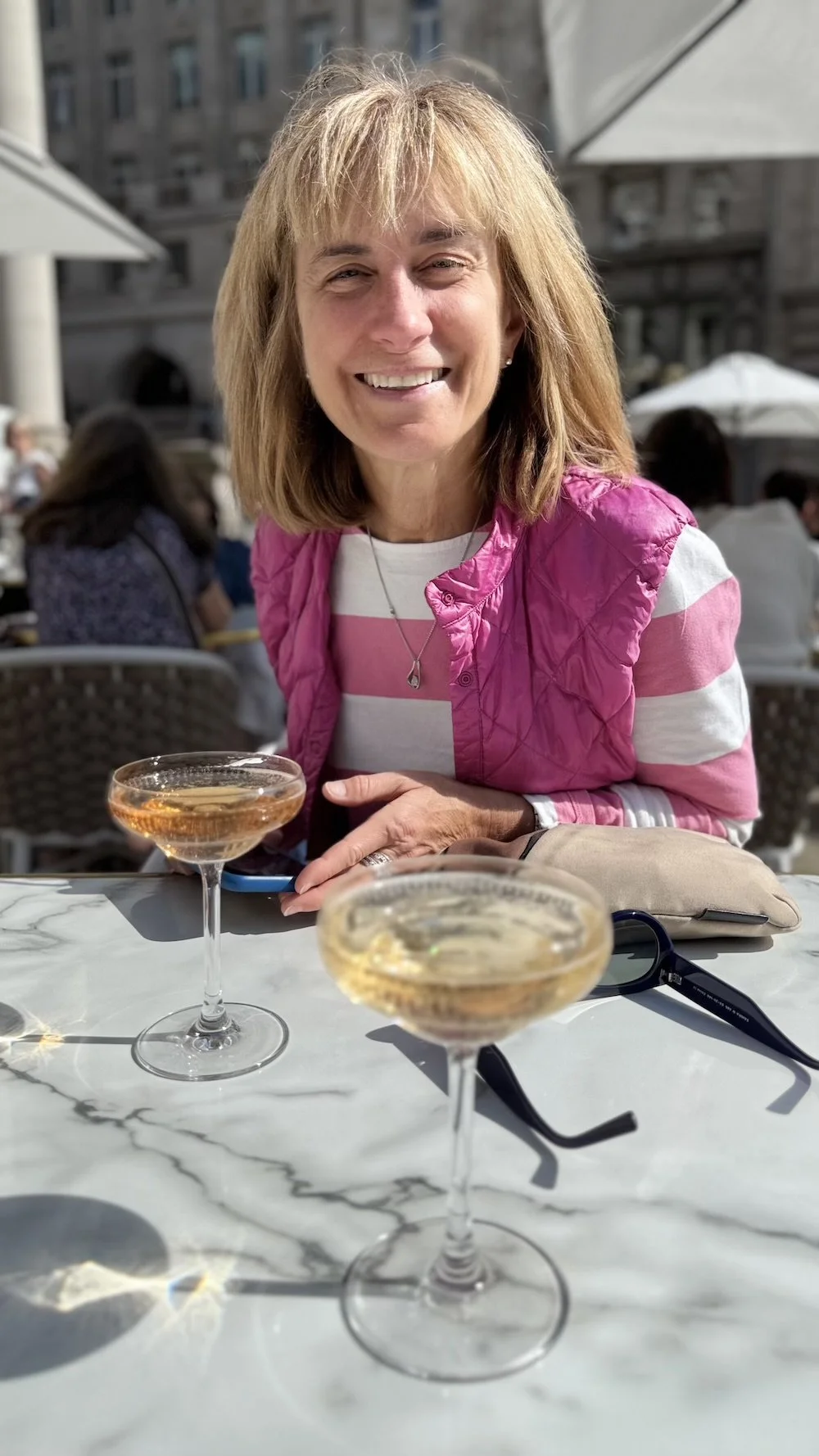 A woman in a pink vest and striped shirt smiling at an outdoor cafe with two glasses of champagne on a white marble table.