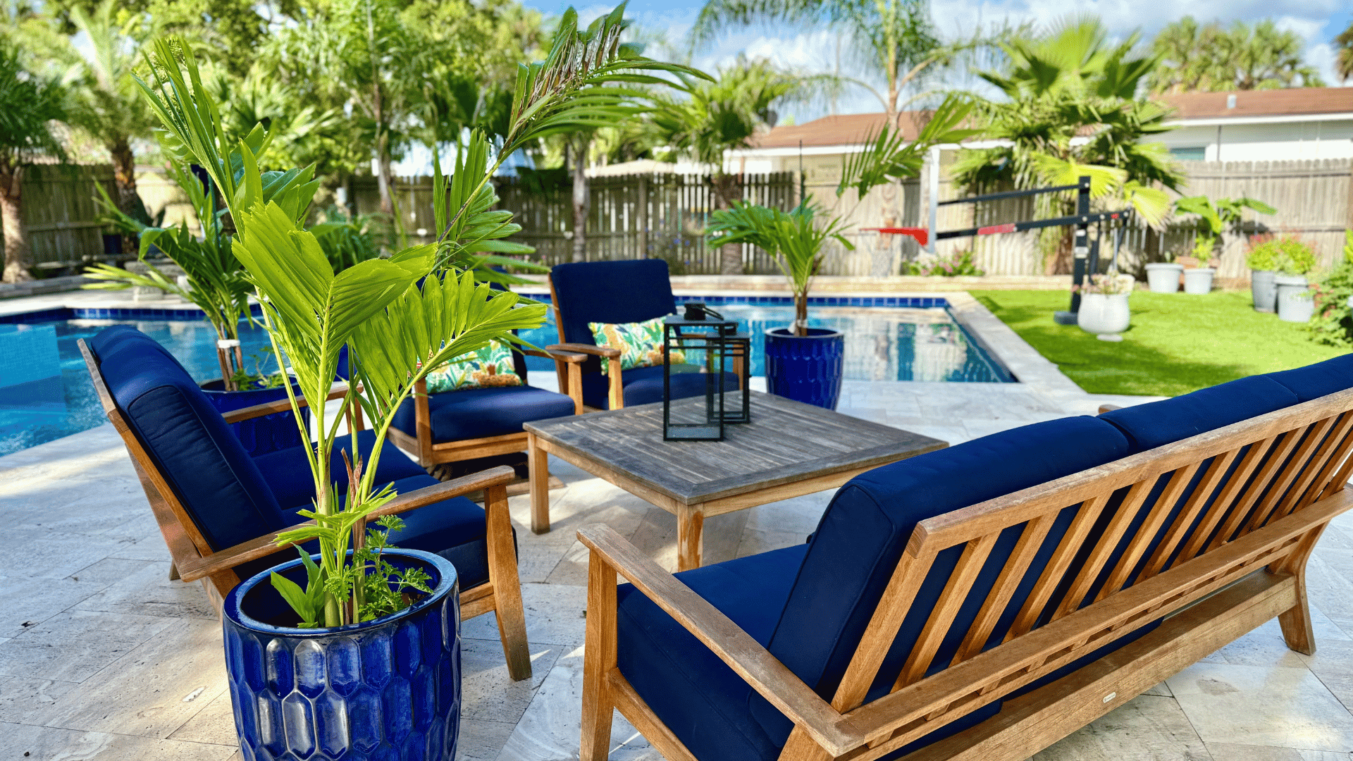 Backyard with a swimming pool, outdoor furniture with blue cushions, potted plants, and green trees.