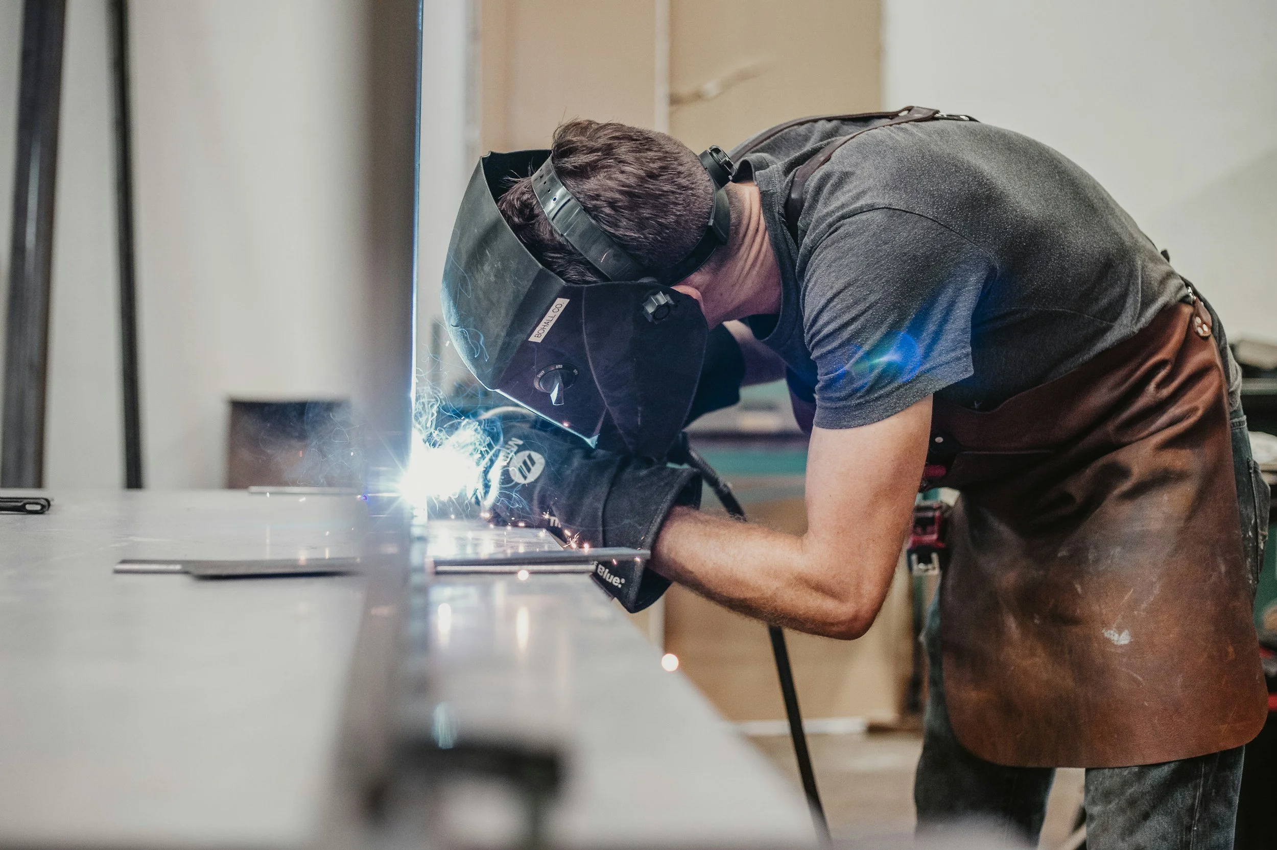 A man welding metal while wearing protective gear, including a face shield and gloves, in a workshop.
