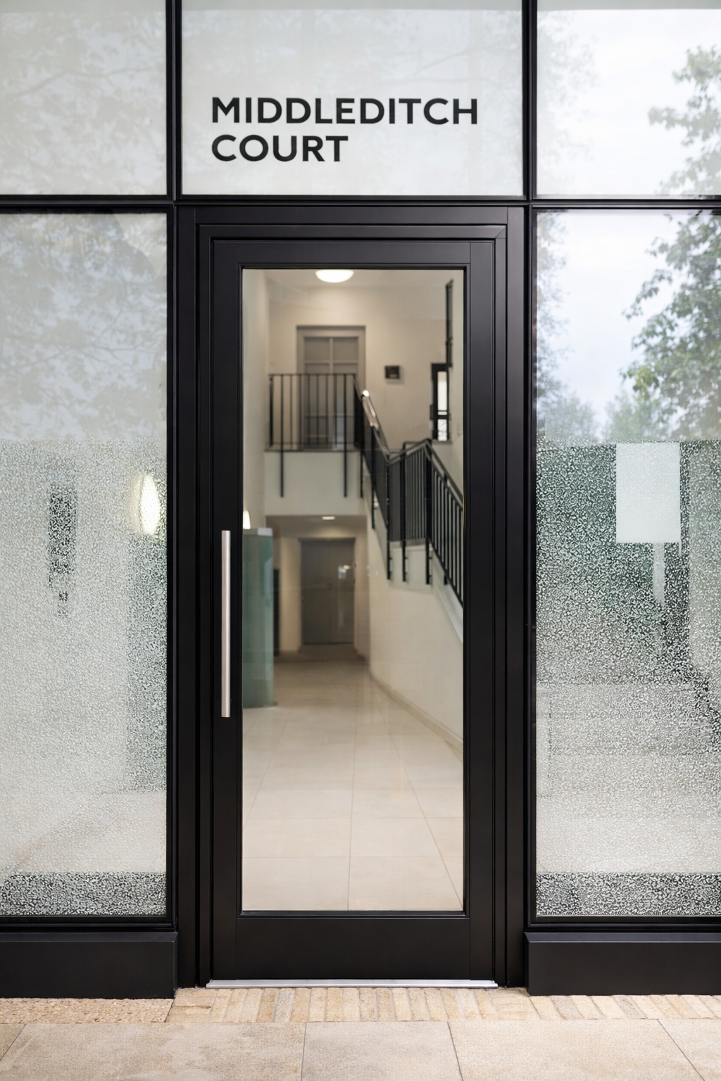 Glass door with black frame leading into a modern, well-lit building interior, with stairs visible through the door, and frosted glass on either side.