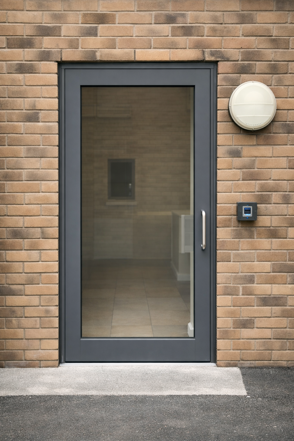 Glass door with a metal handle on a brick wall. There is a white circular device and a black security keypad or intercom on the wall beside the door.