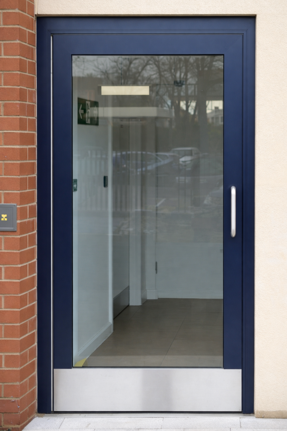 Photo of a closed glass door with a blue frame and silver handle, leading into a building with a brick wall on the left and a beige wall on the right, with reflections of trees and parked cars on the glass.