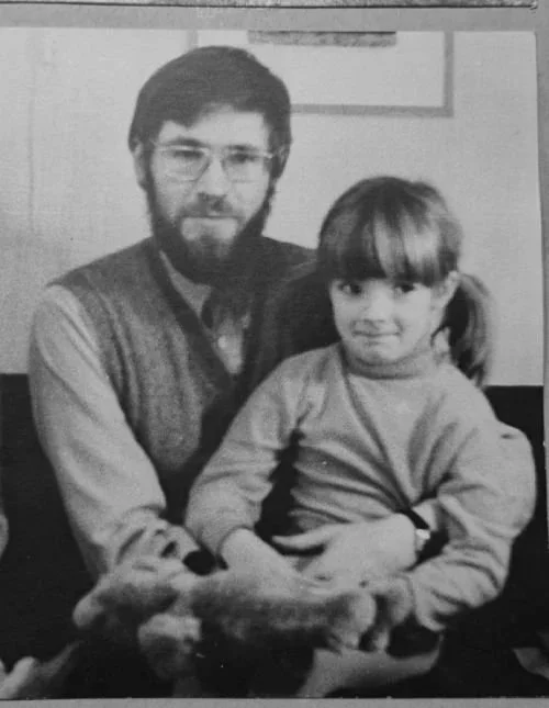 Svart-hvitt fotografi av en far med hår, skjegg og briller sitter sammen med sin datter i en lenestol. Hun har pannelugg og langt, og holder en bamse i hendene.