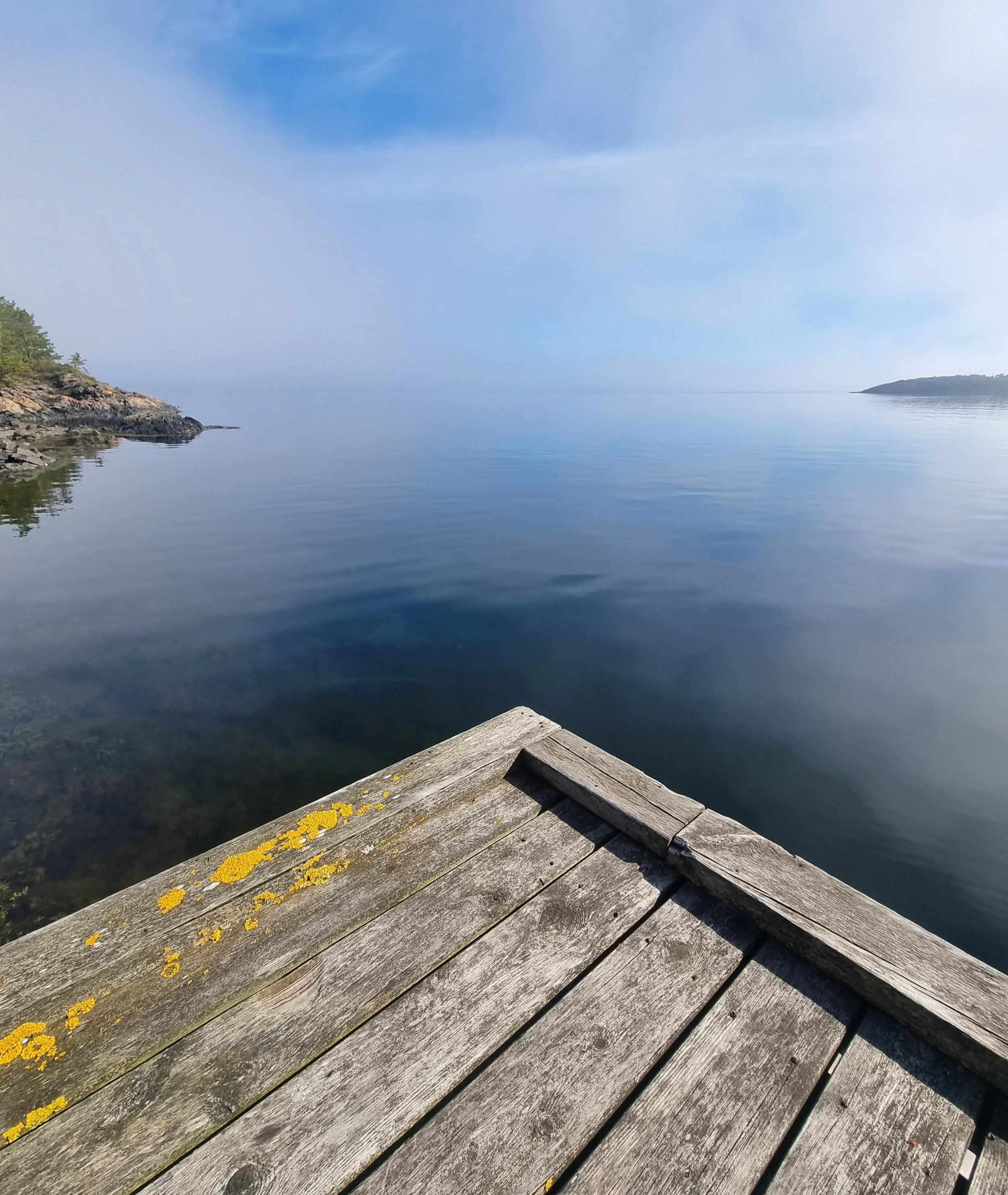 Utsikt fra en bryggekant, mot rolig sjø med svaberg på sidene, med land i horisonten under en blå himmel.