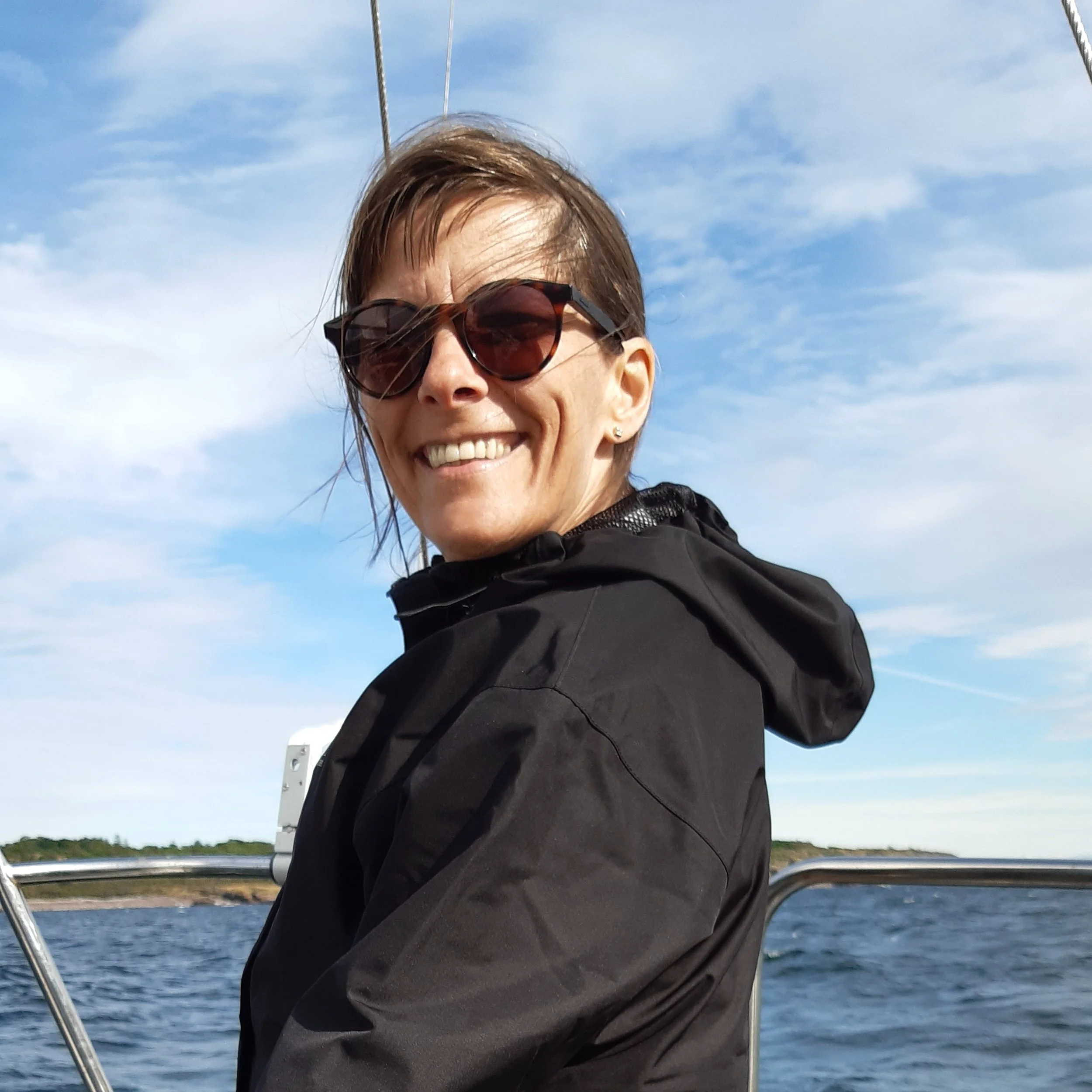 Nærbilde av en terapeuten, med brunt hår og solbriller, smilende, på en båttur på vannet med klar blå himmel og skyer i bakgrunnen.