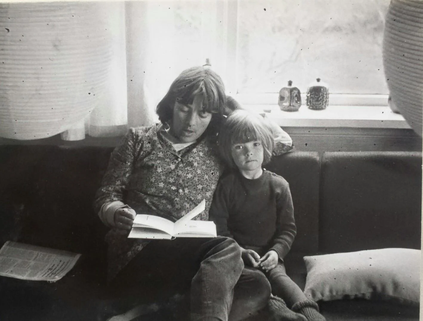 Svart-hvitt fotografi av en mot og et barn som sitter på en sofa. Moren leser en bok, mens barnet ser rett mot kamera. Det er en vindusåpning bak dem med keramikk-figurer i vinduskarmen, og lyse gardiner henger på sidene.
