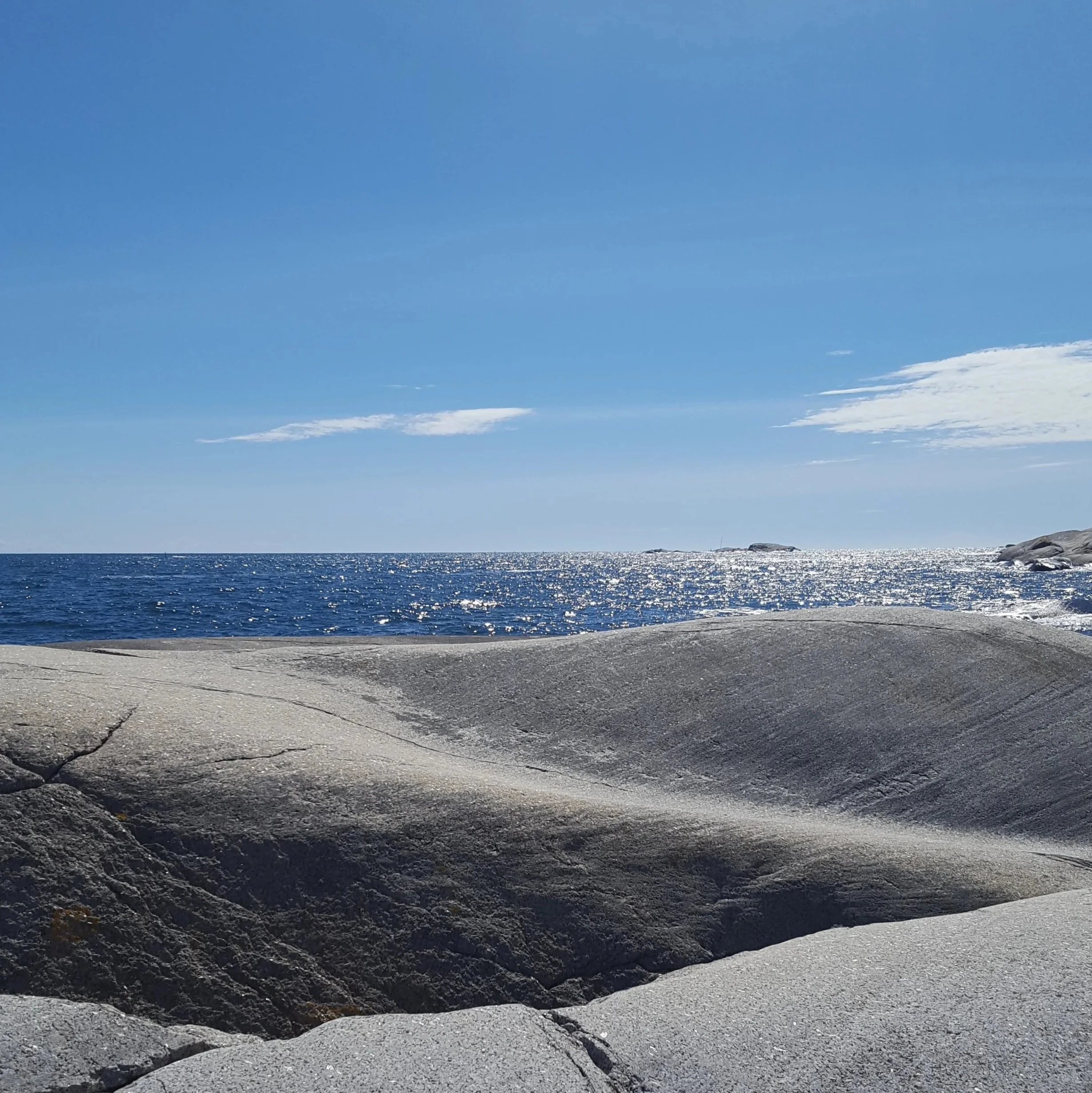Kystlinje med myke svaberg, i bakgr.unnen mørkeblått rolig hav og blå himmel med spredte skyer