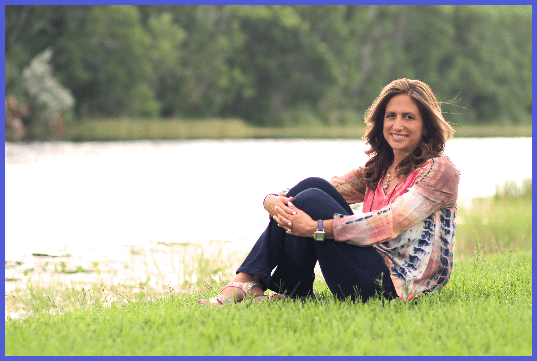 A woman sitting on grass beside a body of water with trees in the background, smiling at the camera.