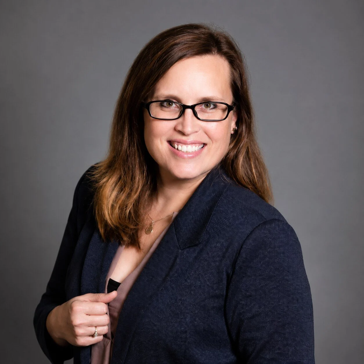 Portrait of a woman with long brown hair, glasses, smiling, dressed in a navy blazer and light-colored blouse, against a neutral gray background.
