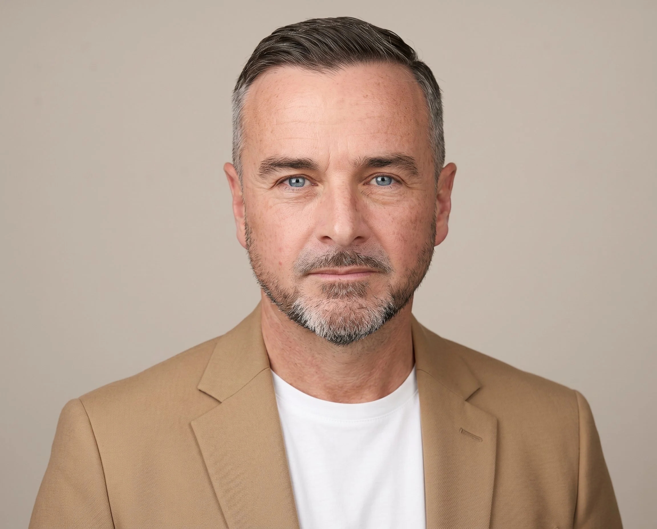 A white man with blue eyes in beige blazer and white t-shirt