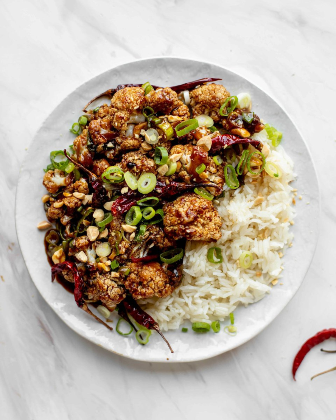 Plate of Chinese-style crispy cauliflower with green onions, dried red chilies, crushed peanuts, served with white rice.