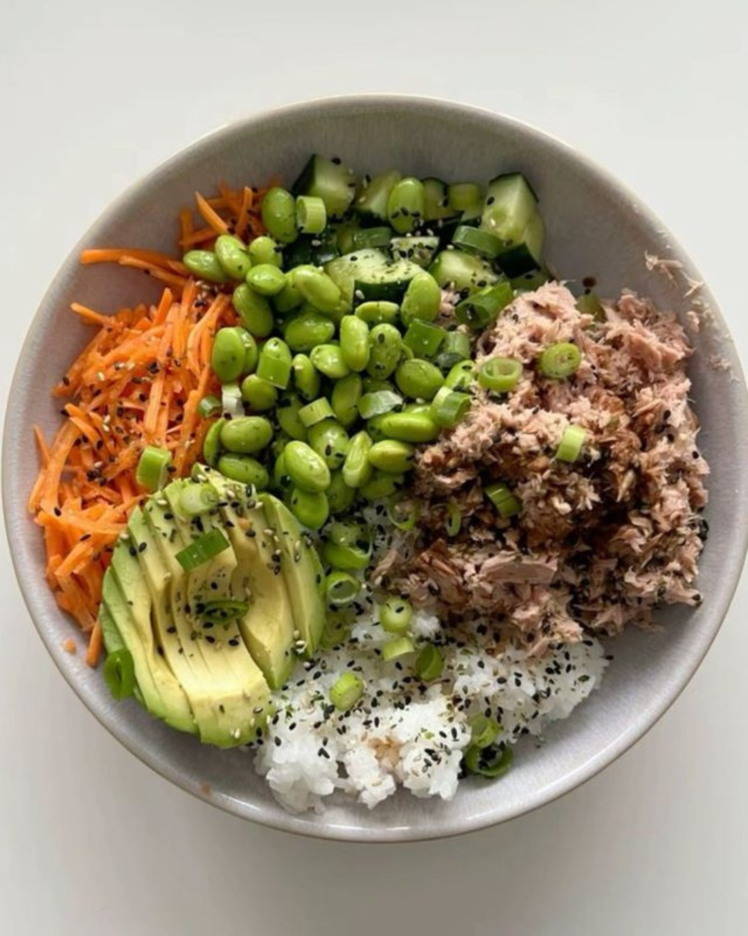 Bowl of poke with rice, avocado, shredded tuna, chopped cucumbers, shredded carrots, edamame, and green onions, garnished with black sesame seeds.