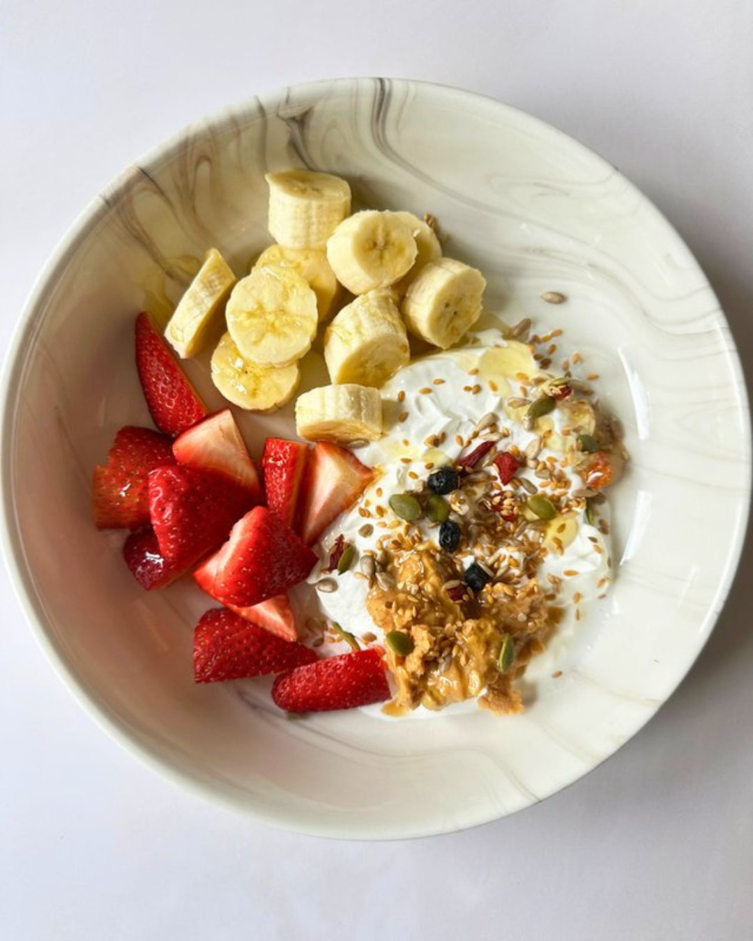Bowl of sliced bananas, strawberries, and yogurt topped with granola, seeds, and dried fruit.