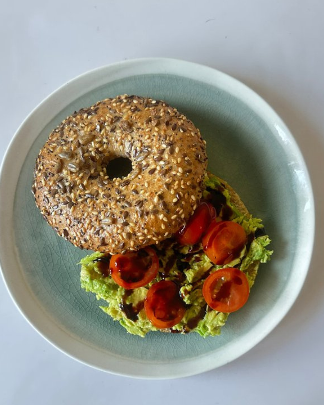 Bagel with sesame and flax seeds served with lettuce, cherry tomatoes, and balsamic glaze on a light blue plate.