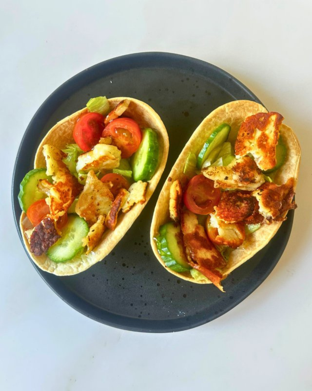 Two tacos on a black plate with tomatoes, cucumbers, and lettuce