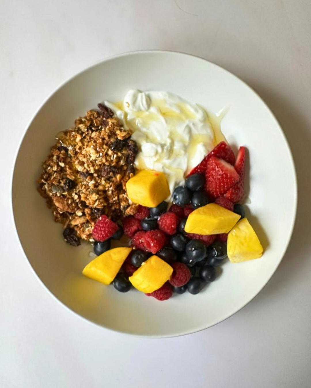 Bowl of yogurt with granola and fresh mixed berries, including strawberries, blueberries, and raspberries, garnished with pieces of ripe mango.