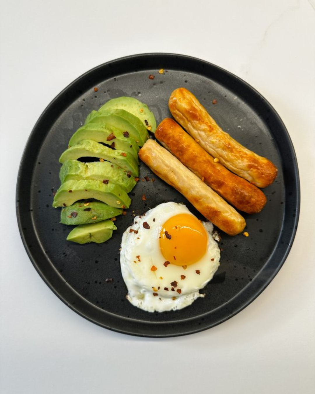 Plate with sliced avocado, four cooked sausages, fried egg topped with red pepper flakes.