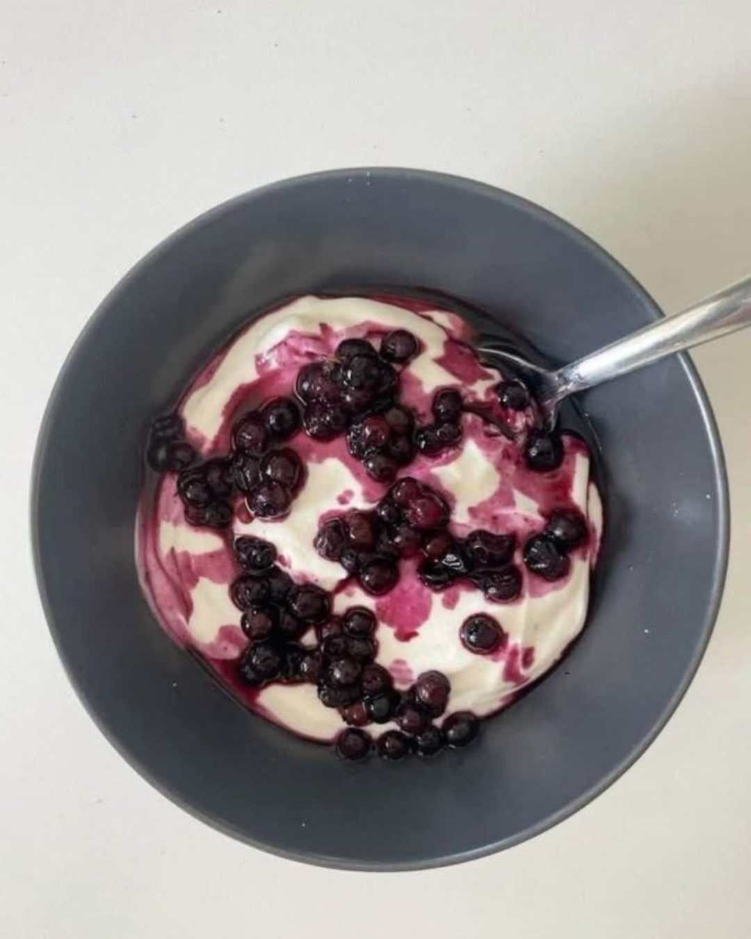 A black bowl containing vanilla yogurt topped with dark blueberries and a berry sauce, with a spoon inside.