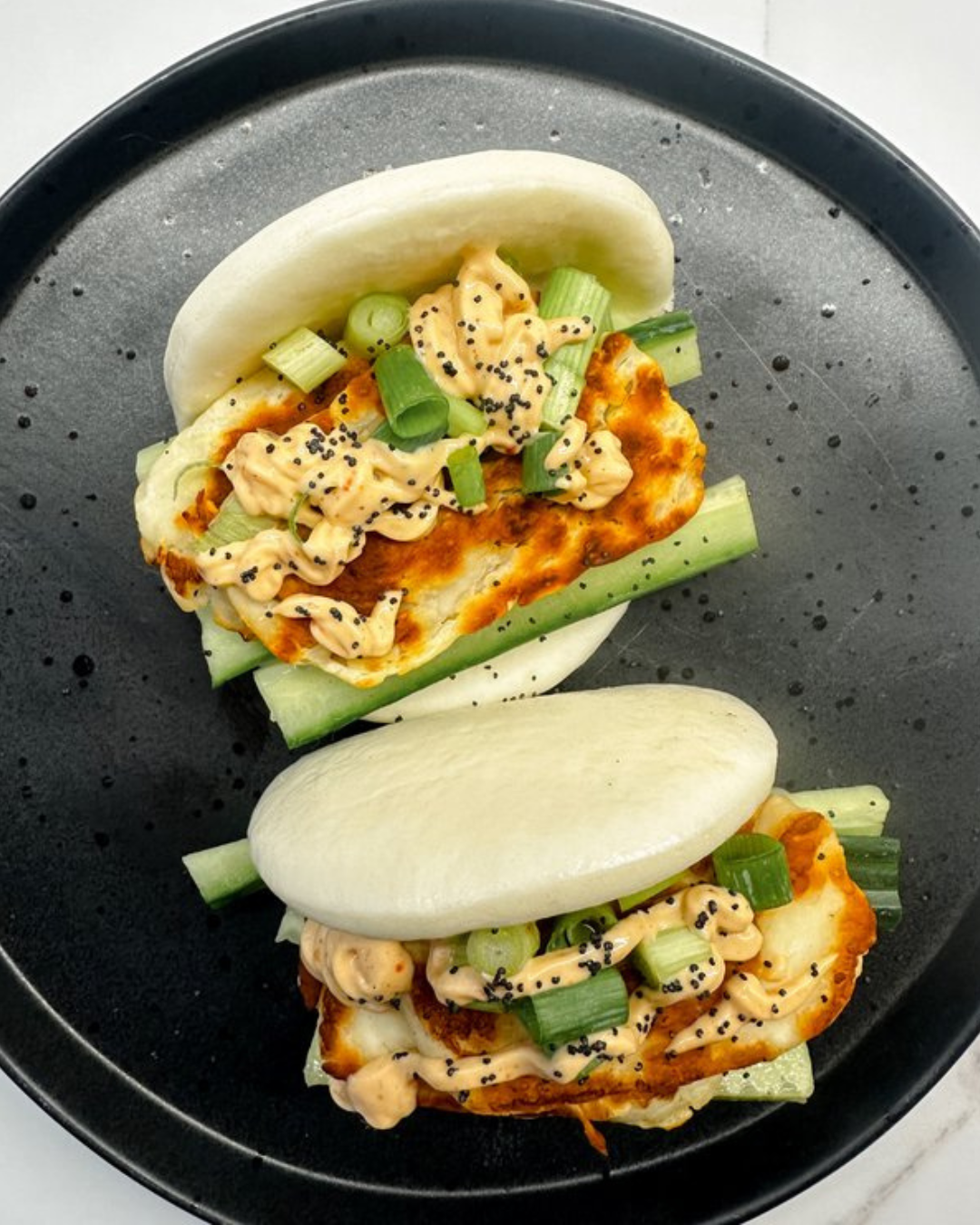 Two steamed bao buns filled with fried chicken, sliced green onions, cucumber, creamy sauce, and black sesame seeds on a dark plate.