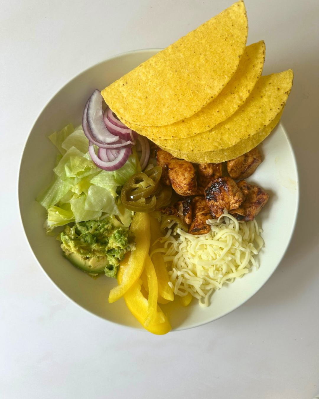 Bowl of Mexican food with nacho chips, grilled chicken, shredded cheese, lettuce, avocado, yellow bell peppers, sliced red onions, and jalapeño peppers.