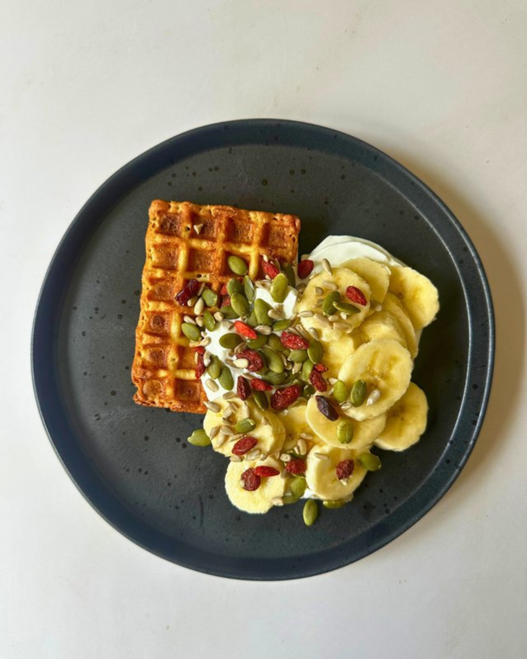 A black plate holds a waffle topped with mixed seeds and dried berries, alongside sliced bananas and a dollop of cream.