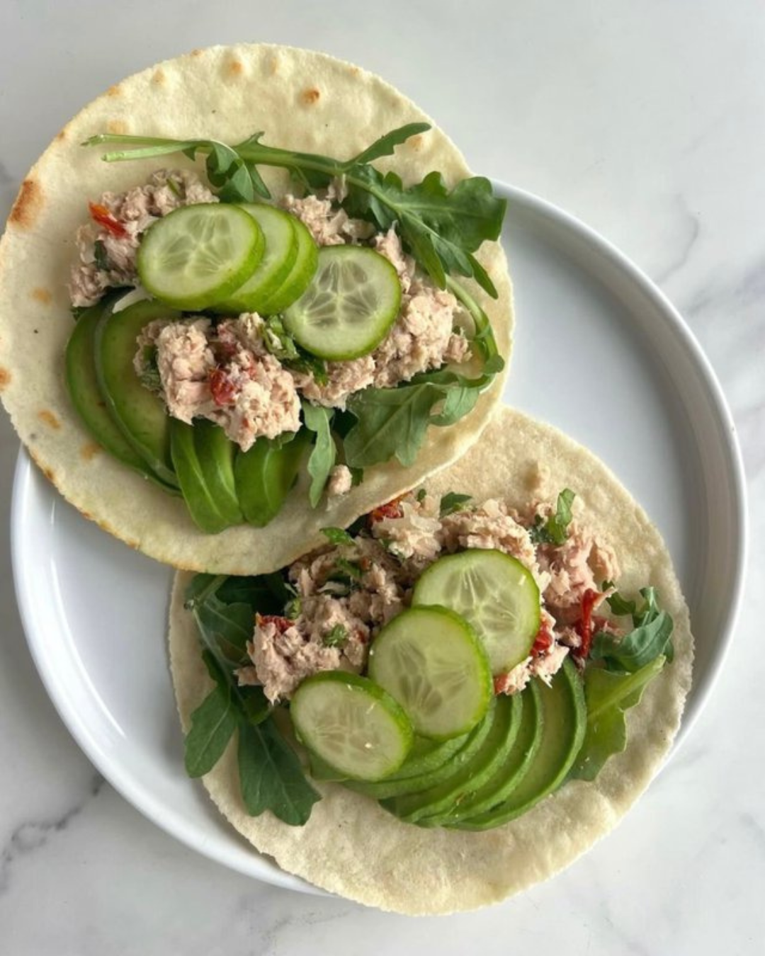 Two tacos filled with tuna salad, cucumber slices, green leafy vegetables, and arugula, served on soft corn tortillas on a white plate.
