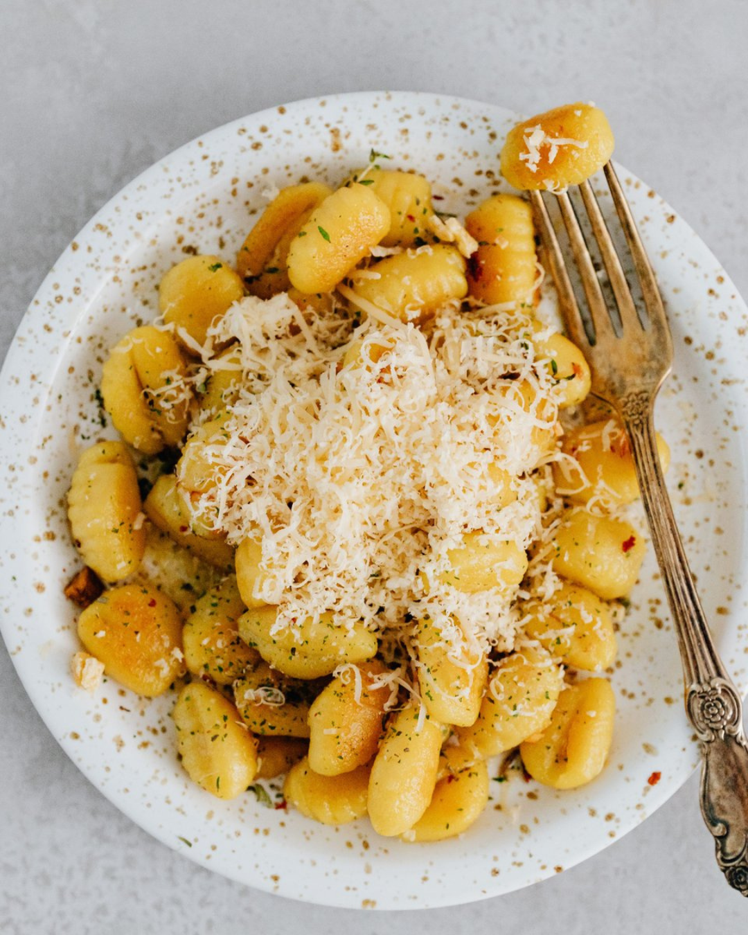 A white plate of gnocchi with shredded cheese and herbs, with a fork resting on the side, on a light gray surface.