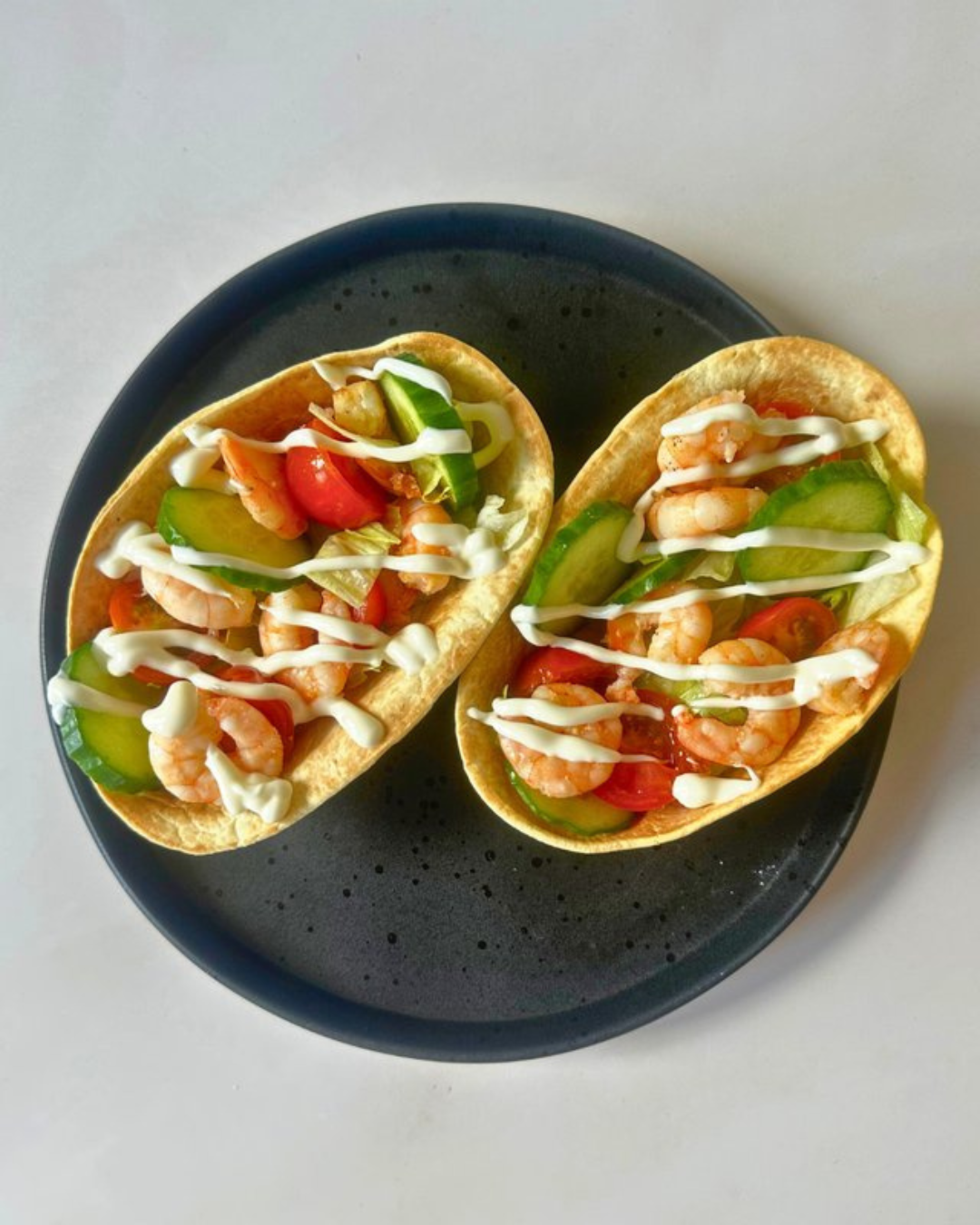Two shrimp tacos on a black plate with chopped cucumbers, cherry tomatoes, and mayonnaise drizzle.