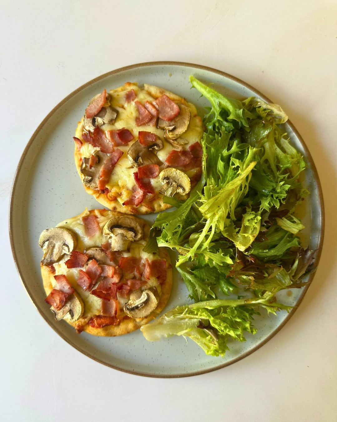 Plate with two small pizzas topped with mushrooms, bacon, and bits of ham, accompanied by a mixed green salad.