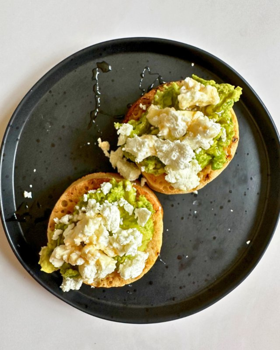 Two open-faced sandwiches with mashed avocado, crumbled feta cheese, on toasted bread on a black plate.