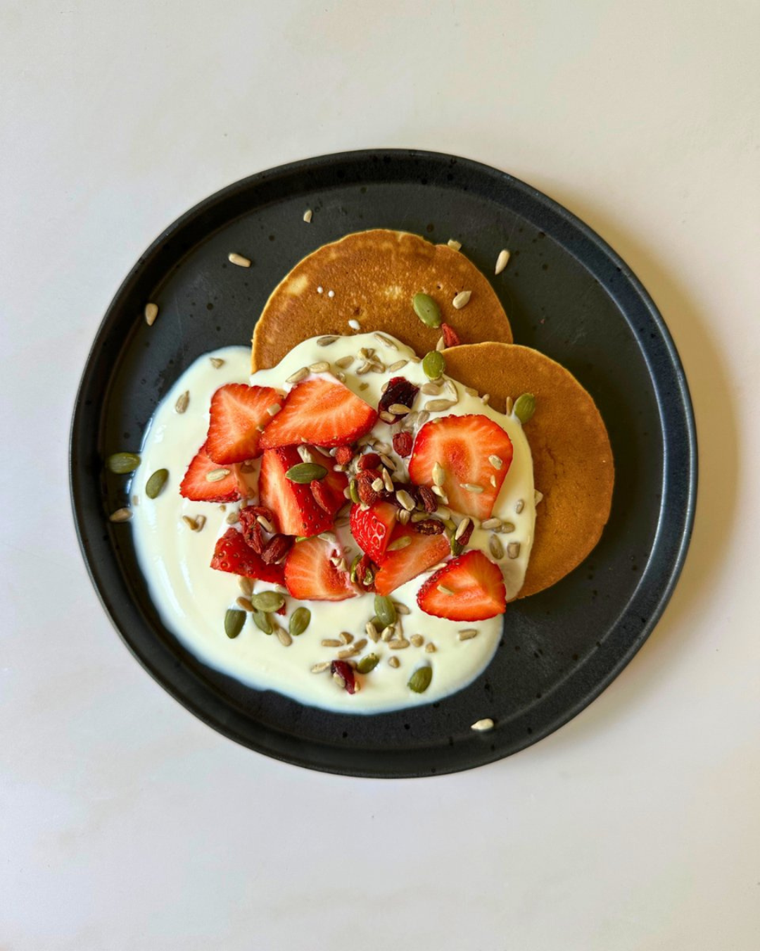 Plate with two golden-brown pancakes topped with strawberries, yogurt, mixed seeds, and a small amount of cream, on a black plate with a white background.