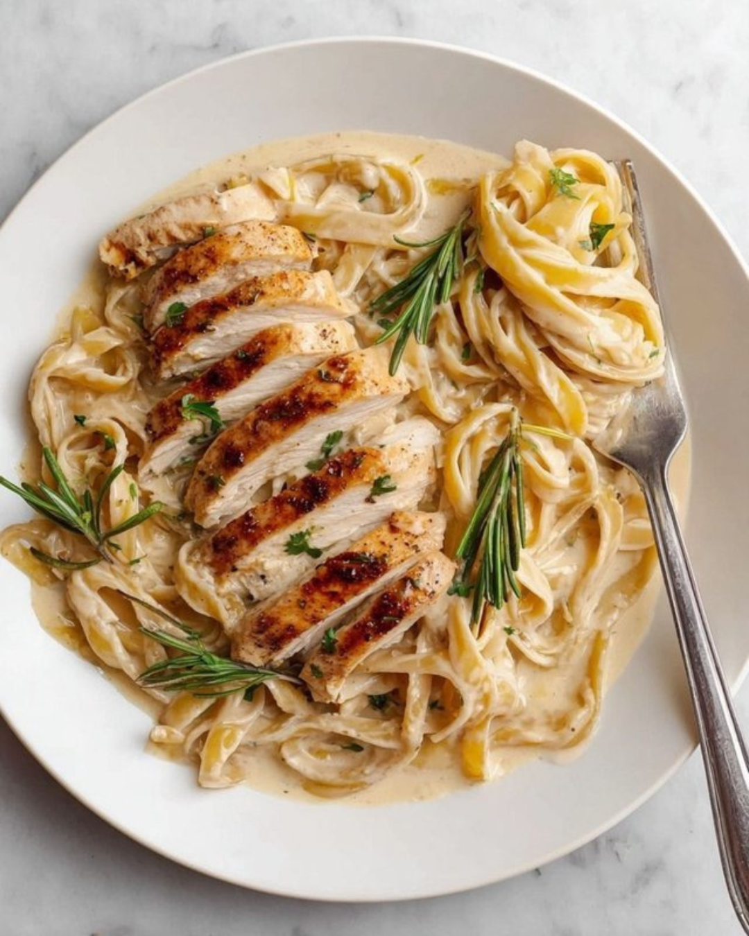 Creamy pasta with grilled chicken slices garnished with rosemary and parsley on a white plate.