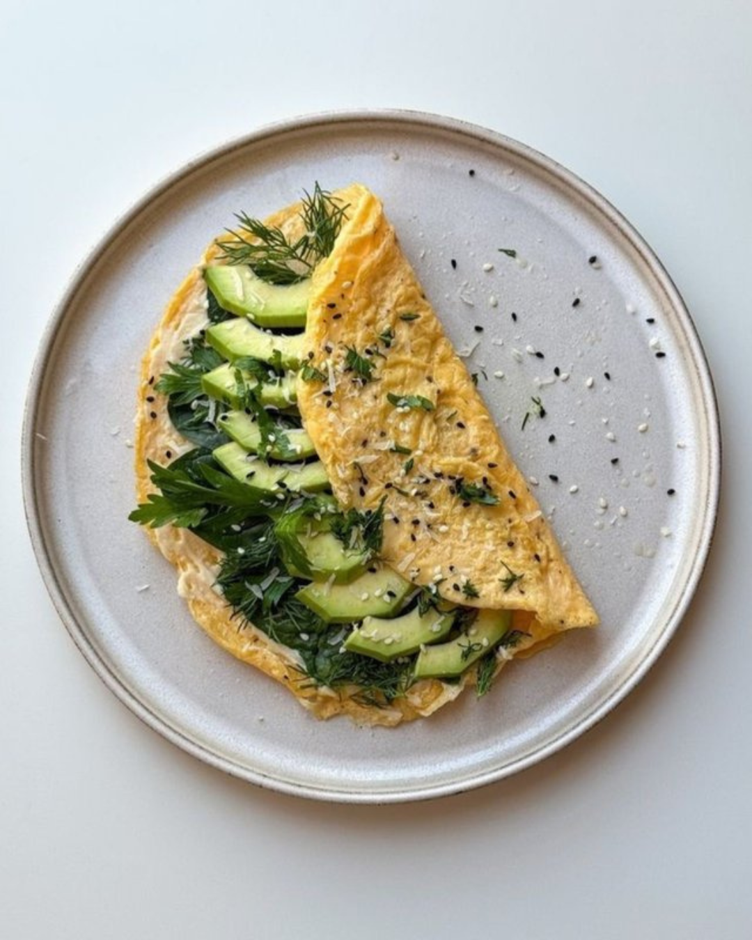 Omelette filled with sliced cucumbers and fresh herbs, garnished with sesame seeds on a white plate.