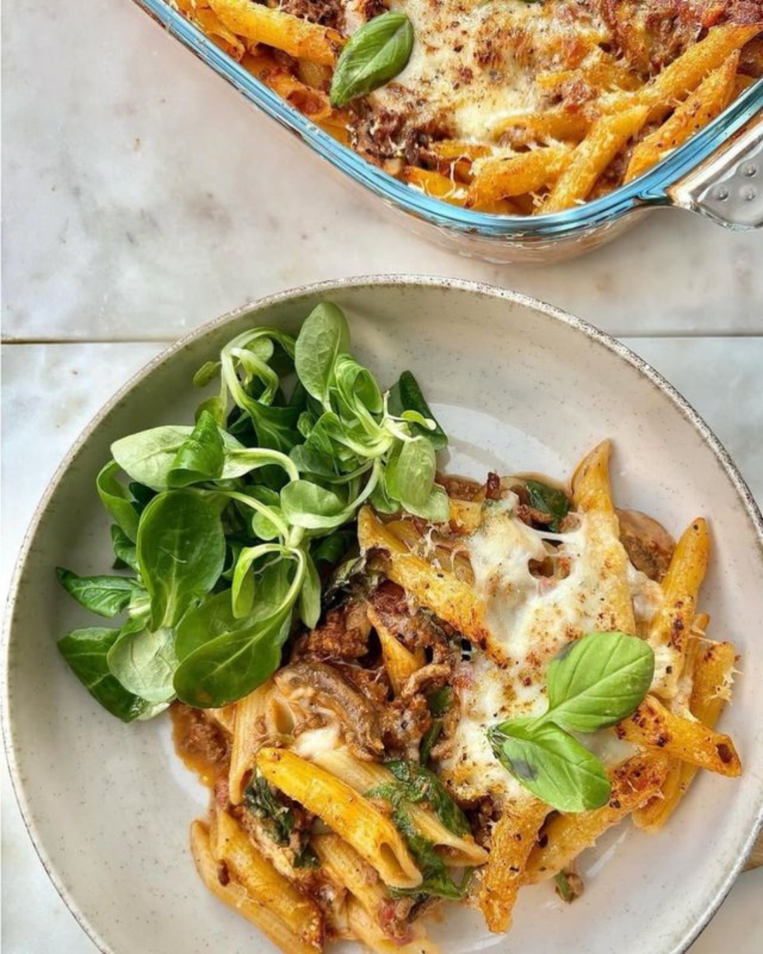 A plate of baked pasta with cheese, meat, and vegetables served with fresh greens, and a glass baking dish containing more pasta in the background.