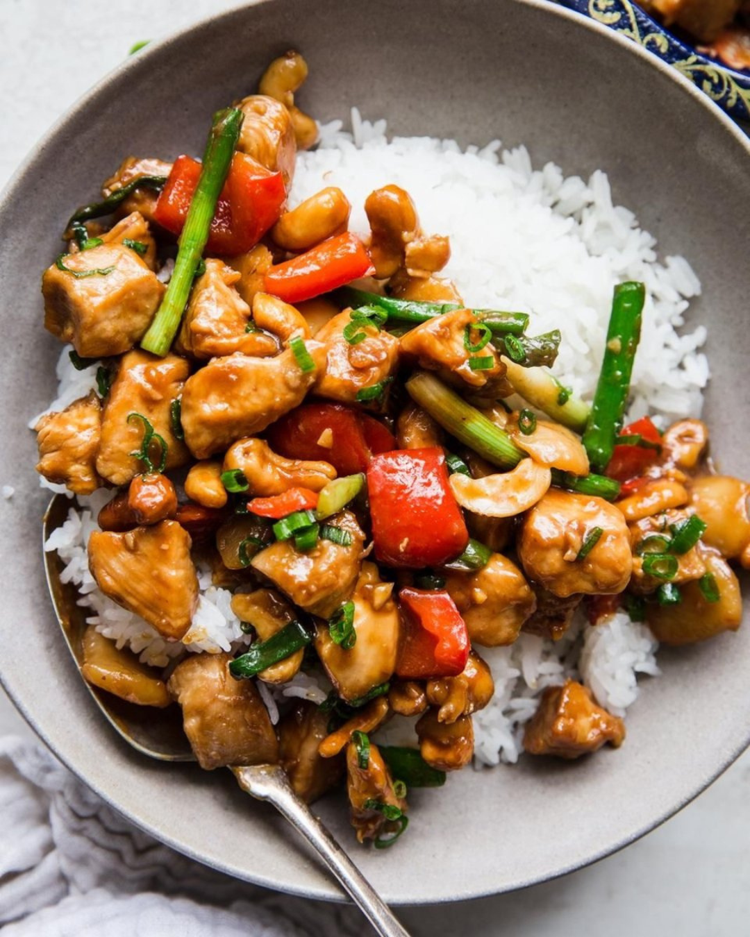 A bowl of white rice topped with stir-fried chicken and vegetables including red bell peppers, green onions, and cashews in a brown sauce.