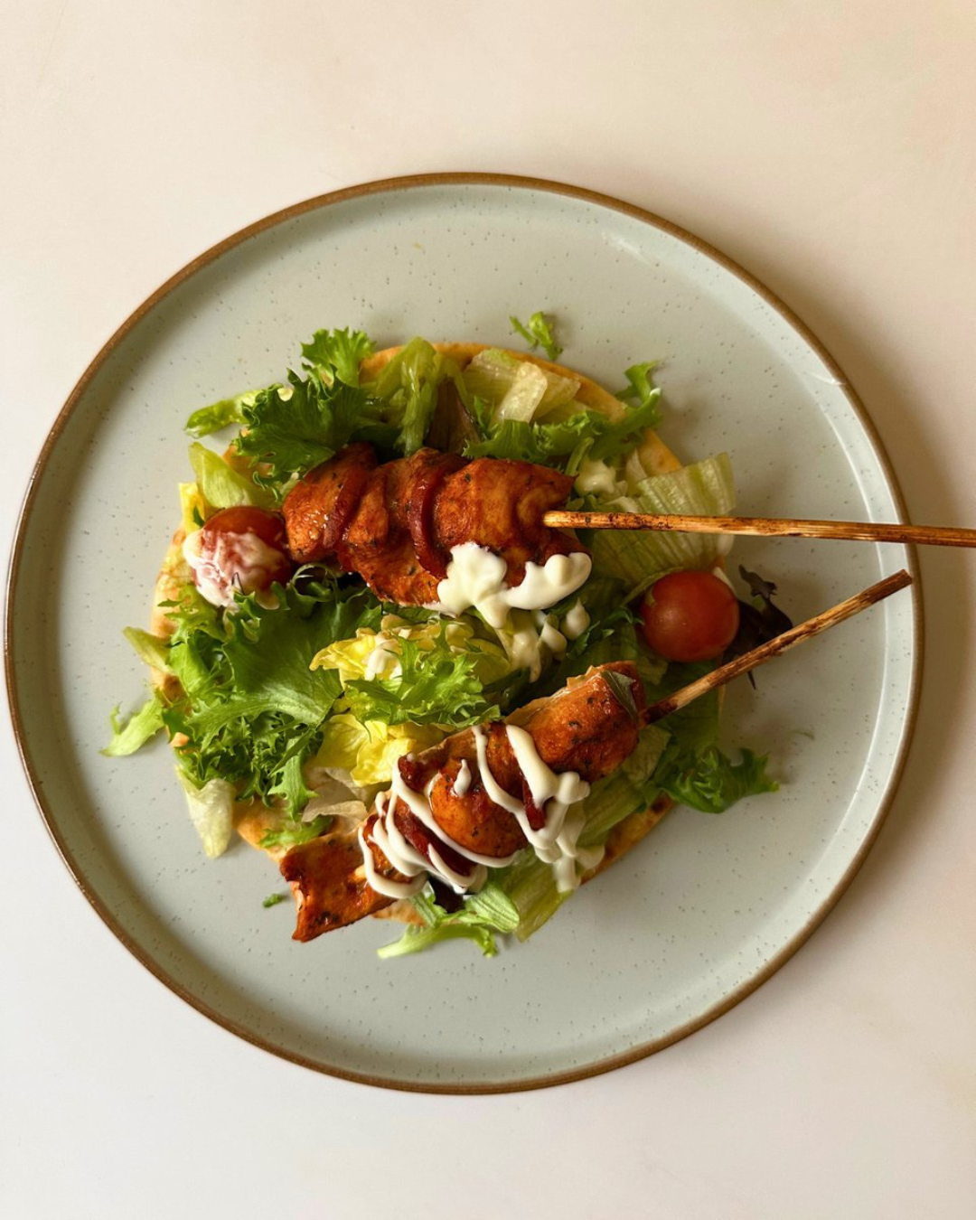 Plate of grilled chicken skewers on a bed of lettuce with cherry tomatoes, drizzled with mayonnaise and ketchup.