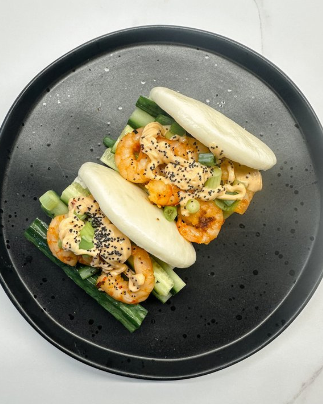 Two Asian steamed buns filled with shrimp, topped with chopped green onions, black sesame seeds, and a creamy sauce, served with cucumber and lettuce on a black plate.