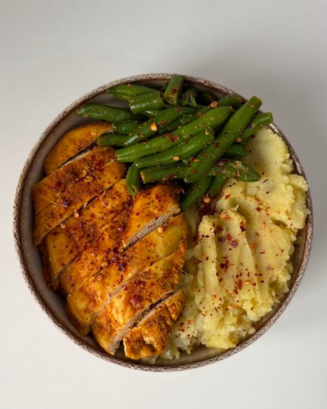 A bowl of mashed potatoes sprinkled with red pepper flakes, green beans with chili flakes, and roasted sliced chicken breast with seasoning.