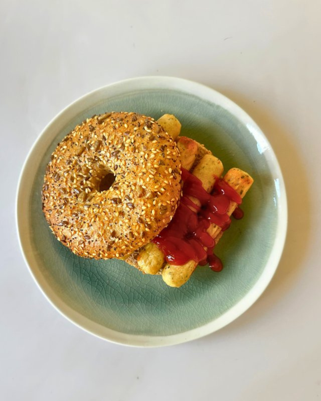 A bagel topped with sesame seeds, served with grilled plantains drizzled with ketchup in a green bowl.