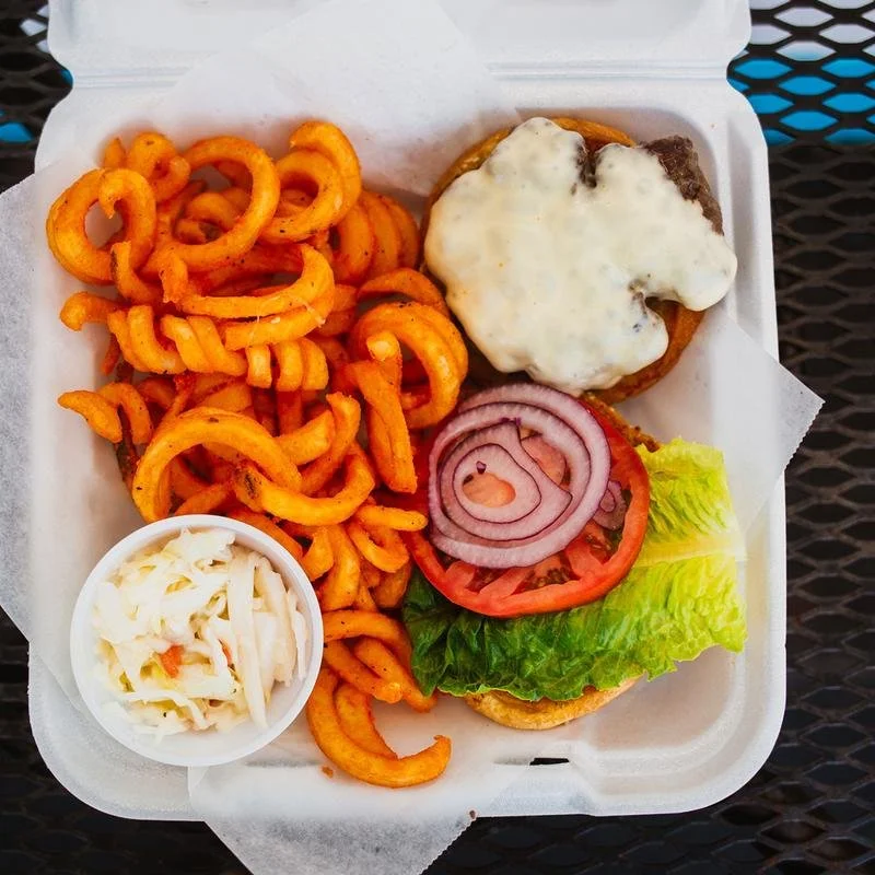 A burger with lettuce, tomato, red onion, and a beef patty topped with white sauce, served with curly fries and a side of coleslaw in a styrofoam takeout container.