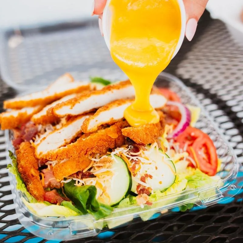 A plastic container of salad topped with fried chicken strips, sliced cucumbers, tomatoes, shredded cheese, and bacon, with yellow salad dressing being poured on top.