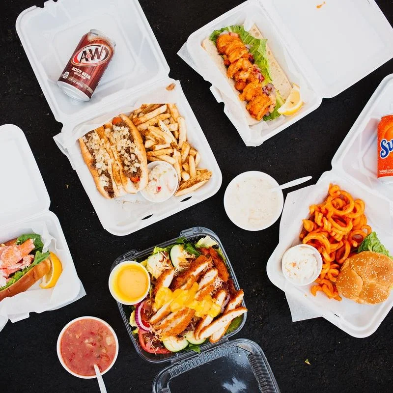A variety of takeout food containers on a black table, including a hot dog with toppings and fries, a seafood salad, a chicken sandwich, mozzarella sticks with dipping sauce, curly fries with a burger, a cup of salads, and a bottle of root beer.