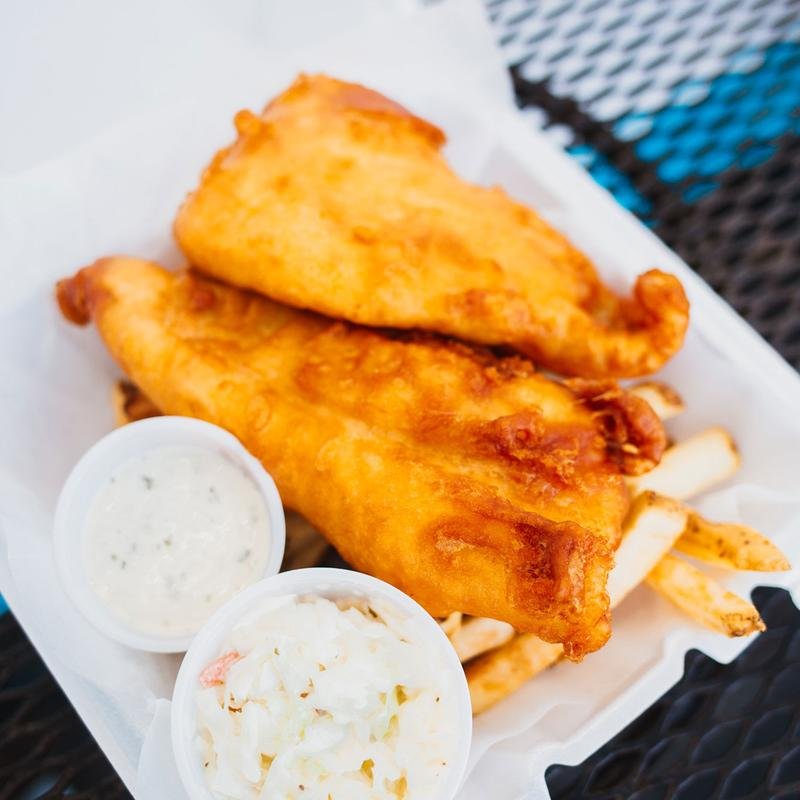 Two pieces of fried fish fillets with a side of French fries, accompanied by two cups of tartar sauce and coleslaw, placed on a paper-lined basket on a dark mesh table.