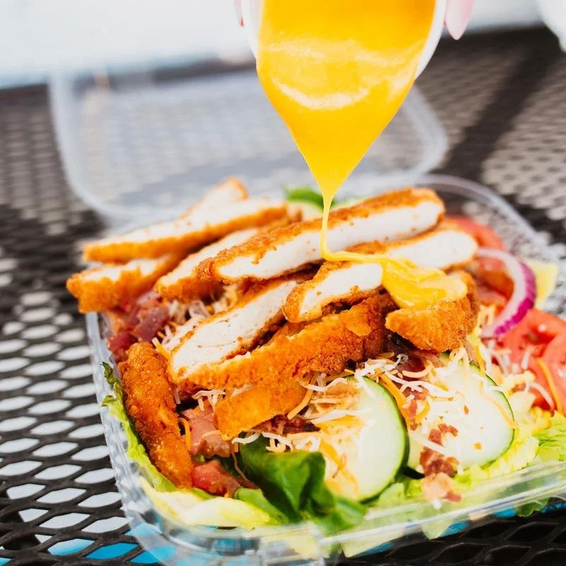 Salad topped with breaded chicken strips, shredded cheese, cucumber slices, tomato slices, and red onion rings with egg yolk being poured over.