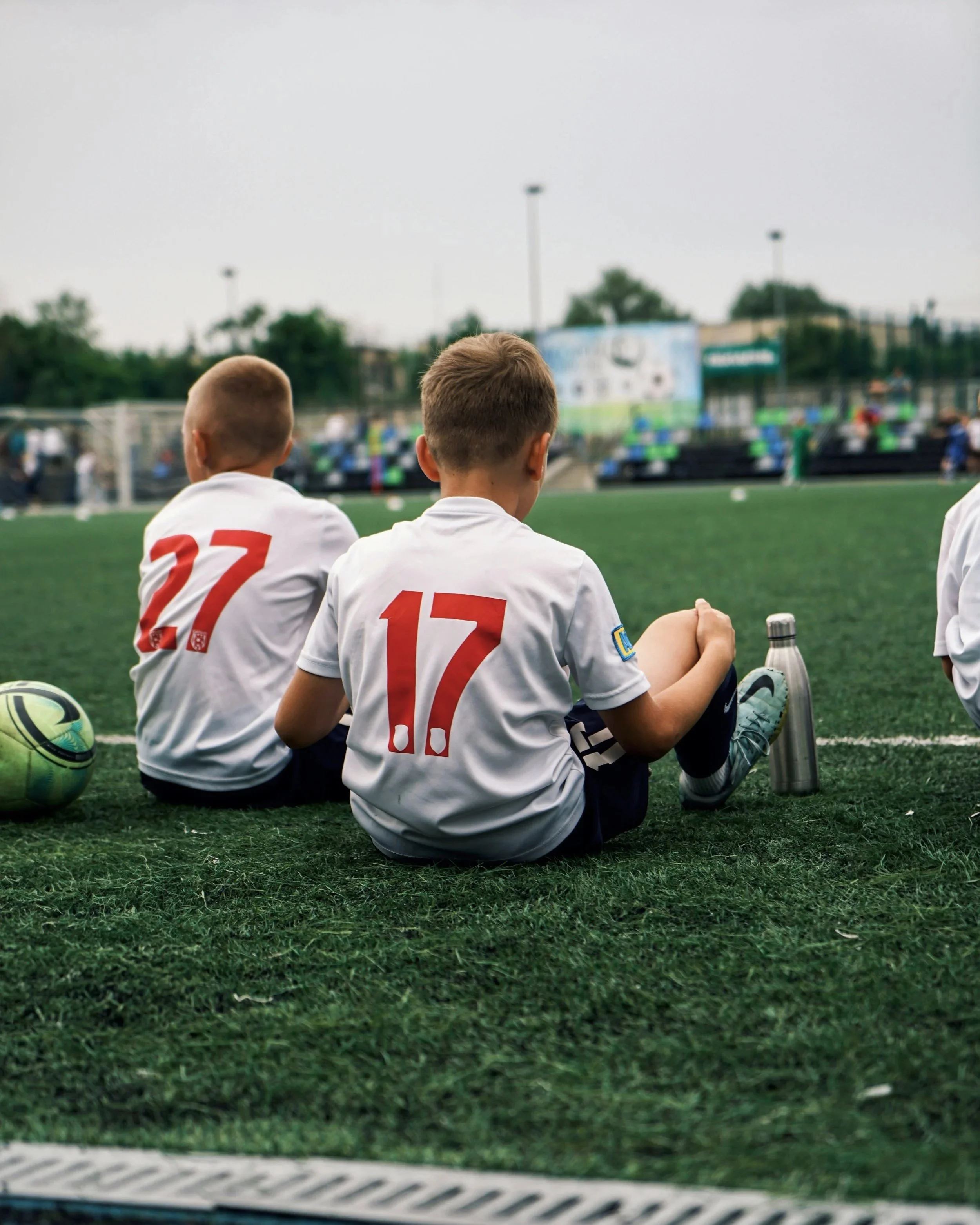 Two young boys with short hair, wearing white sports jerseys numbered 27 and 17, sitting on a soccer field bench watching a game. A soccer ball and a water bottle are beside them, with a blurred soccer match happening on the field in the background.