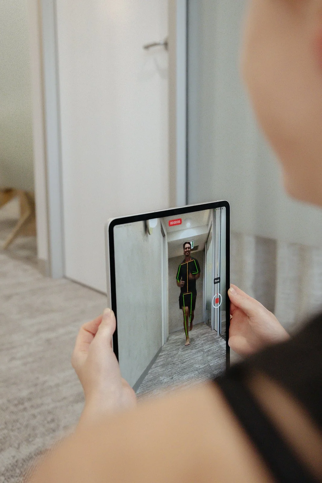 A podiatrist from Peregian Foot Clinic holding a tablet takes a selfie in front of an elevator, with a man in athletic sports wear running in the background.