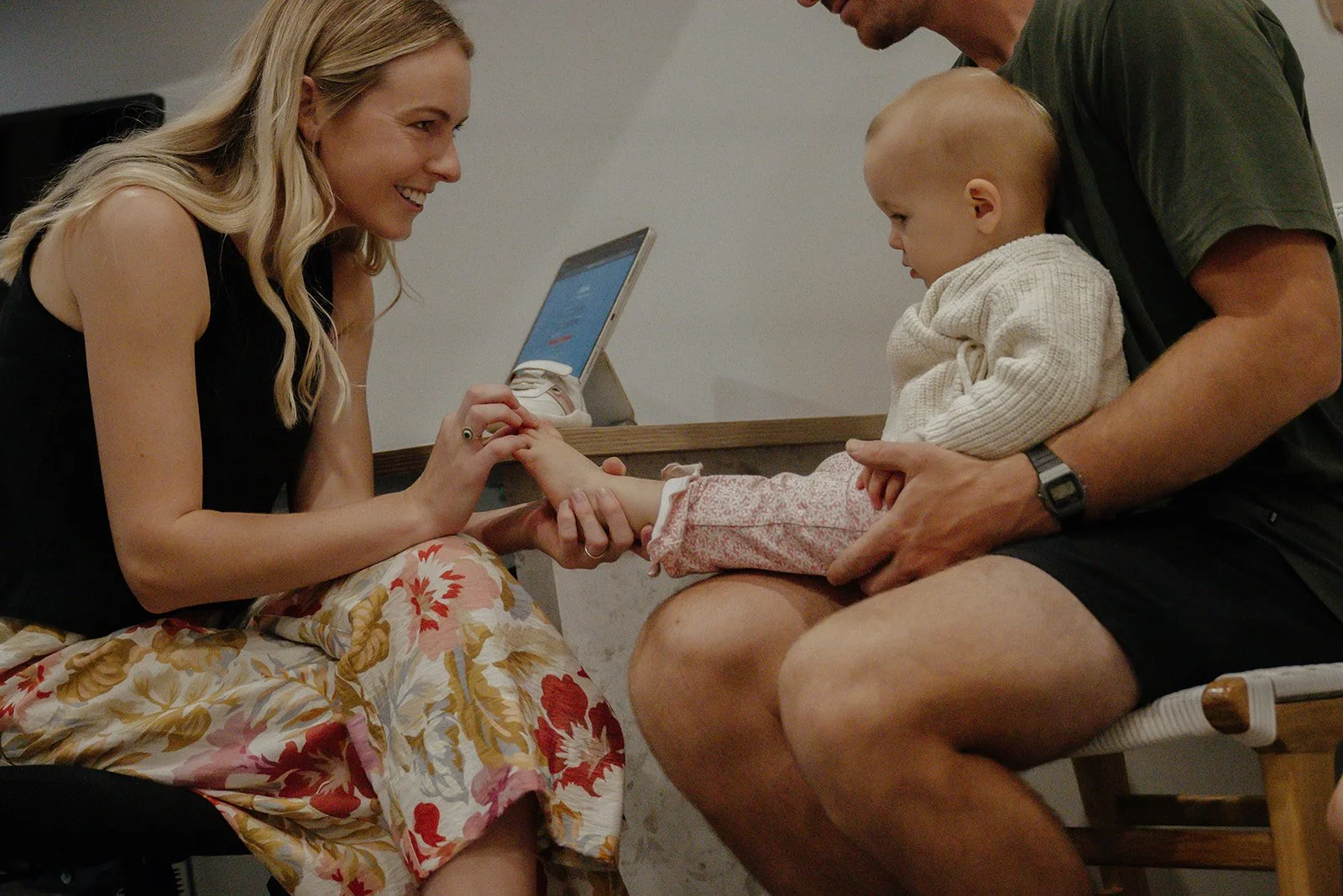 A woman with long blonde hair sitting on the floor, smiling and holding a child's foot, while a man holds the child's leg. The woman appears to be examining or playing with the child's foot, who is sitting on the man's lap. There is a device on the table in the background.