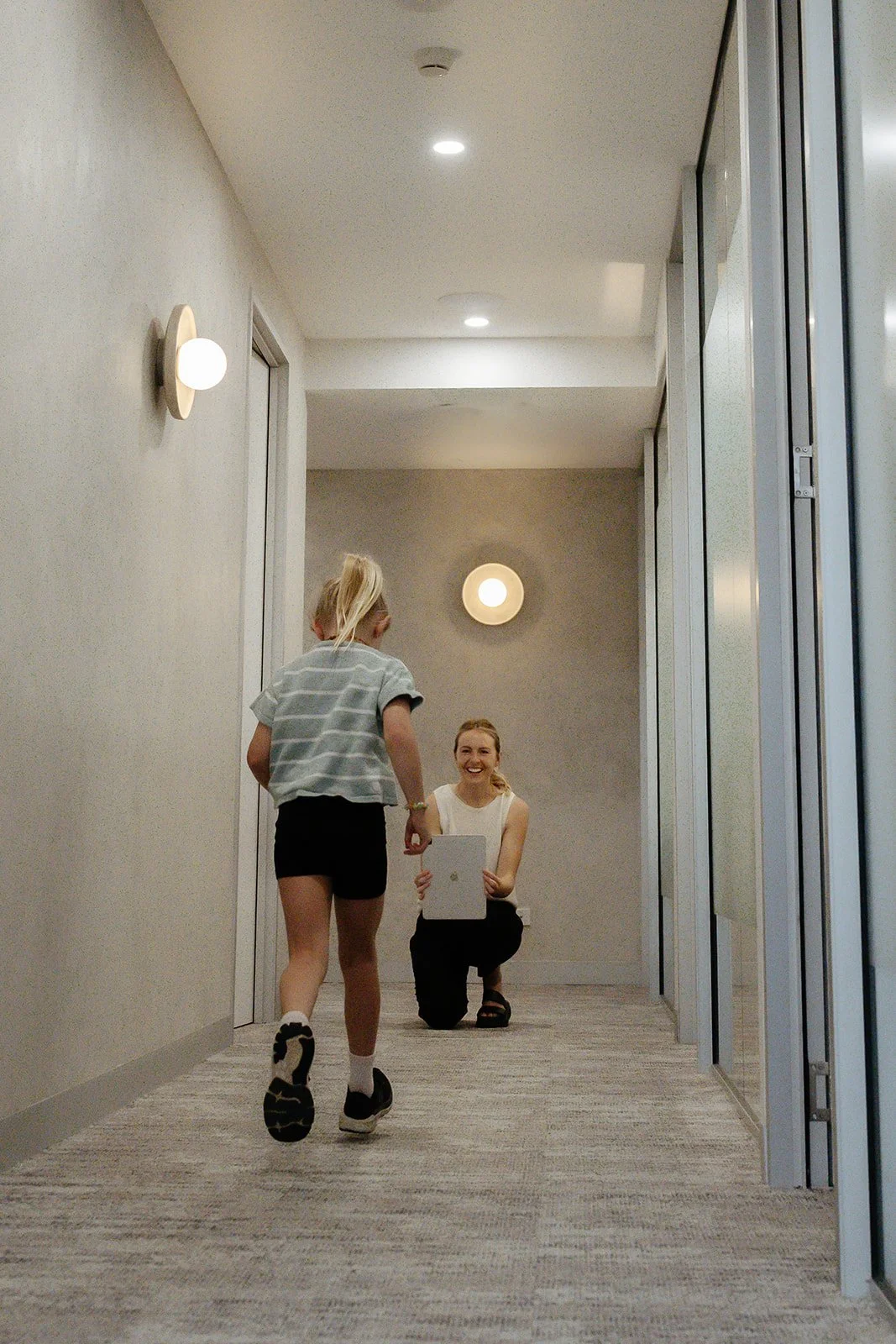 A woman kneeling in a hallway holding a tablet and smiling at a girl running towards her.
